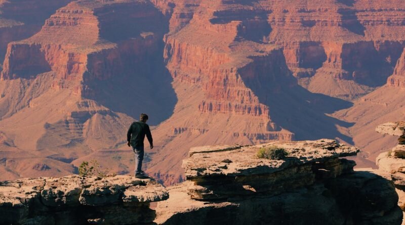A man hikes along the rocky edge of the Grand Canyon under a clear blue sky, capturing the essence of outdoor adventure.
